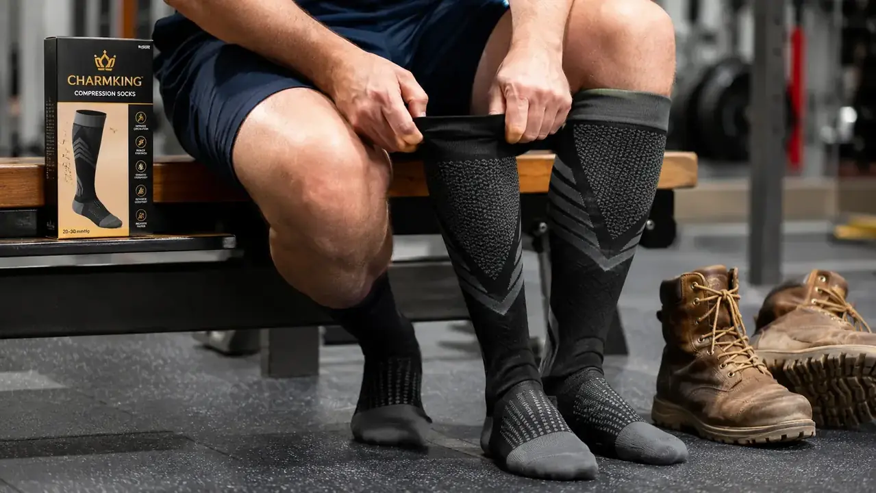 Person pulling on a CHARMKING compression sock while seated on a gym bench, work boots visible on the floor nearby