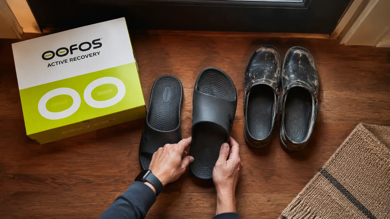 OOFOS OOahh recovery slides on a hardwood floor near a front door, next to a pair of work clogs